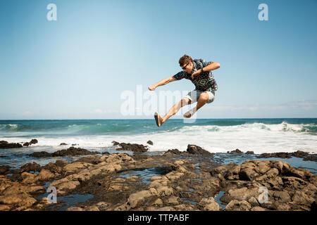 Man jumping on rocky shore against blue sky Stock Photo