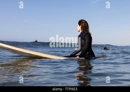Thoughtful female surfer sitting on surfboard in sea against clear sky Stock Photo
