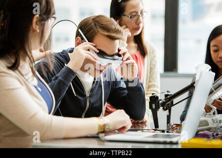 Male student wearing virtual reality glasses while sitting by friends in classroom Stock Photo