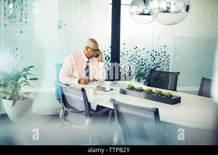 Doctor using mobile phone while writing notes in board room seen through glass Stock Photo