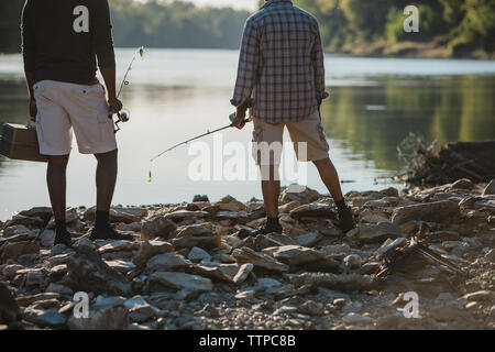 Low section of male friends with fishing rods standing at lakeshore Stock Photo