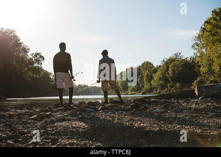 Rear view of friends holding fishing rods while standing at lakeshore against sky Stock Photo