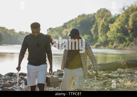 Friends holding fishing rods talking while walking on rocks by lake against sky Stock Photo