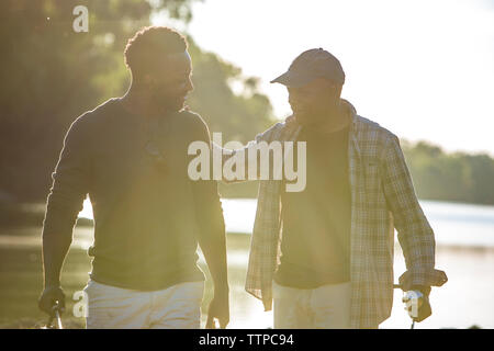 Smiling male friends talking while walking by lake against sky Stock Photo