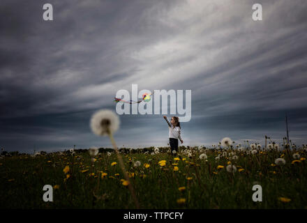 Girl playing with kite while standing on dandelion field against stormy clouds Stock Photo