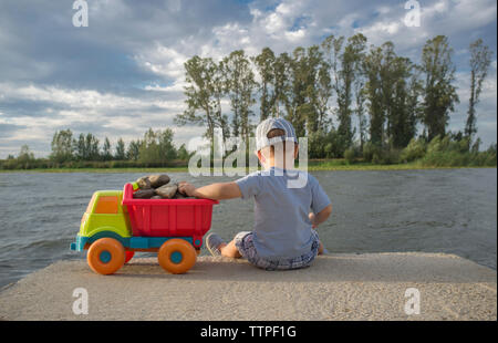 Two year old boy playing at riverside with dump truck. He is sitting over fisher platform throwing stones Stock Photo