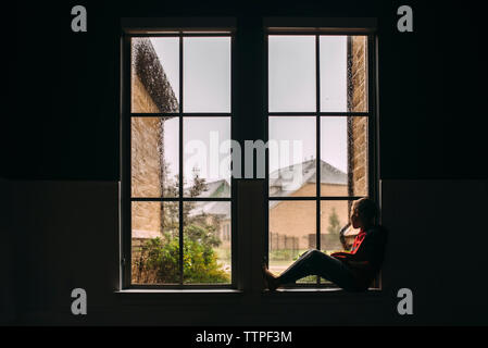 Side view of boy looking through window while sitting at home during rainfall Stock Photo