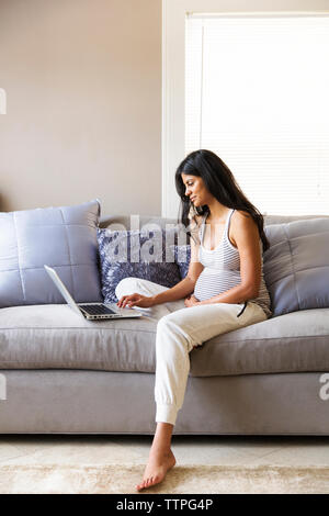 Pregnant woman sitting on sofa using laptop at home Stock Photo