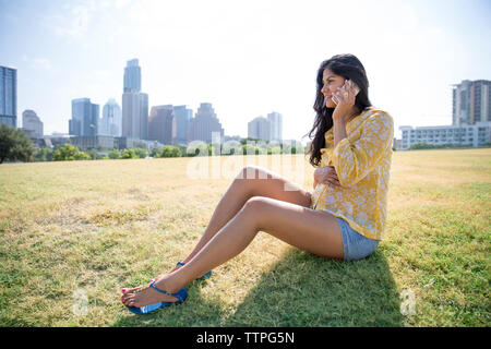 Pregnant woman using phone while sitting on grass against city Stock Photo