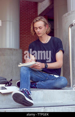 Male student sitting on stairs and writing on notebook Stock Photo