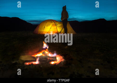 Woman standing by illuminated tent on field against sky during dusk with campfire in foreground Stock Photo