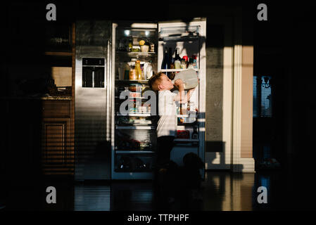 Side view of boy drinking milk from bottle while standing against open refrigerator in darkroom at home Stock Photo