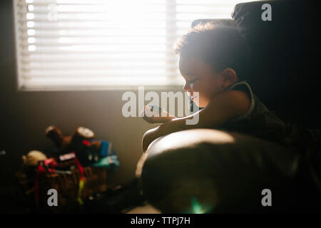 Side view of cute baby boy playing with toy while relaxing on armchair against window at home Stock Photo