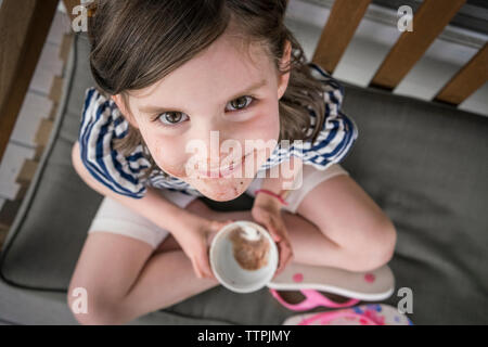 High angle portrait of cute girl with messy mouth eating chocolate while sitting on chair at home Stock Photo