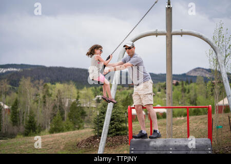 father pushing daughter sitting on chain swing against cloudy sky at park Stock Photo