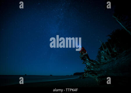 Hiker wearing headlamp sitting on tree trunk at La Push beach against sky at night Stock Photo