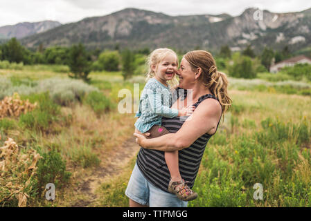 Happy mother carrying cute daughter while standing on grassy landscape against mountains in forest Stock Photo