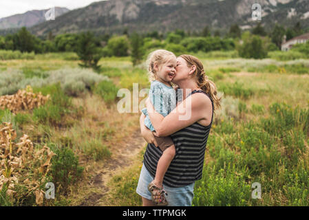 Happy mother kissing cute daughter while standing on grassy landscape against mountains in forest Stock Photo