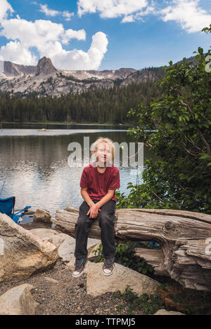 Portrait of smiling boy sitting on tree trunk against lake in forest Stock Photo