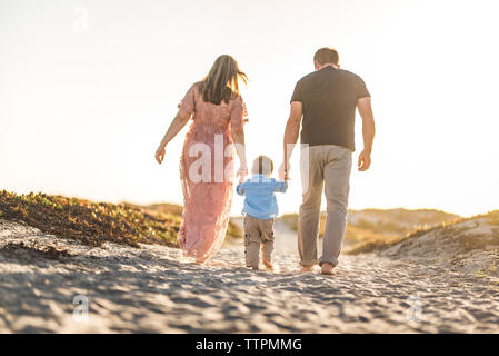 Rear view of parents holding son's hands while walking on sand at beach against clear sky during sunset Stock Photo