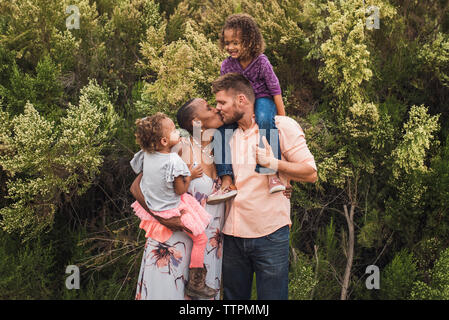 Romantic parents kissing while carrying daughters against trees in park Stock Photo