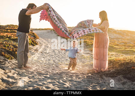 Side view of parents holding blanket over cute son running on sand at beach against clear sky during sunset Stock Photo