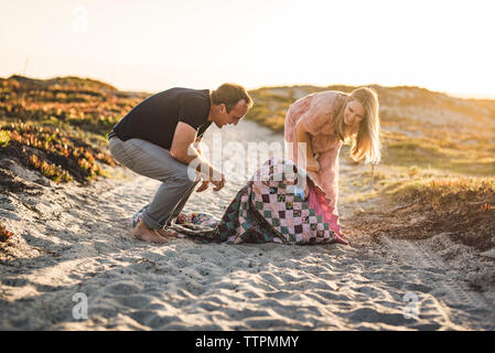Happy parents looking at cute son covered in blanket while sitting on beach against clear sky during sunset Stock Photo