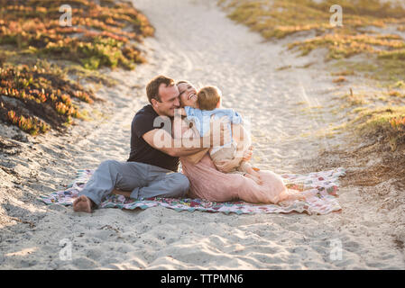 Happy parents embracing son while sitting on sand at beach during sunset