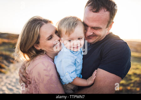 Happy parents embracing son while standing at beach against clear sky during sunset