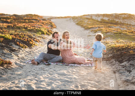 Happy parents gesturing towards son running at beach against clear sky during sunset Stock Photo