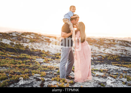 Husband kissing wife while carrying son on shoulders at beach against clear sky during sunset