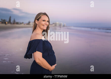Portrait of smiling pregnant woman with hands on stomach standing at beach against sky during sunset Stock Photo