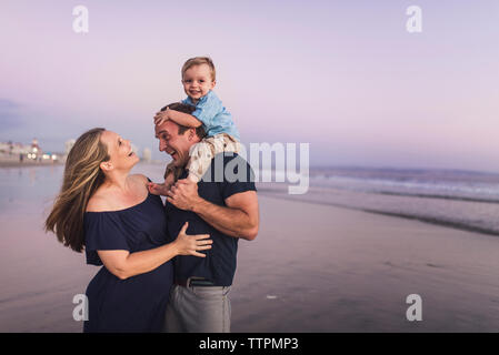Happy husband looking at wife while carrying son on beach against sky during sunset Stock Photo