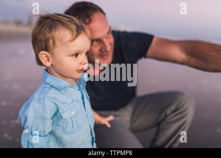 Father and cute son looking away at beach against sky during sunset Stock Photo