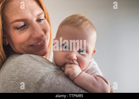 Close-up of mother carrying cute son with fingers in mouth while standing against gray wall at home Stock Photo