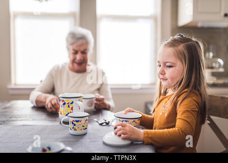 Granddaughter and great grandmother in kitchen playing with tea set Stock Photo