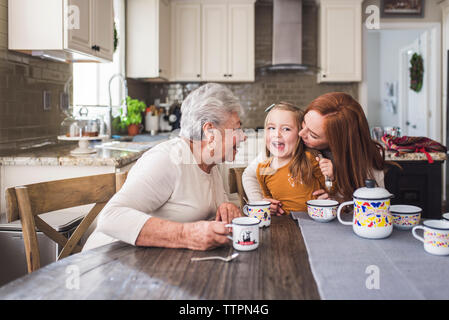 Mother kissing daughter at kitchen table with grandmother looking Stock Photo