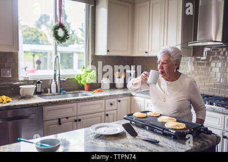 Senior woman drinking coffee while making pancakes for her family Stock Photo