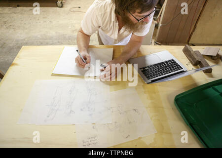 High angle view of carpenter with laptop computer drawing sketch on paper in workshop Stock Photo