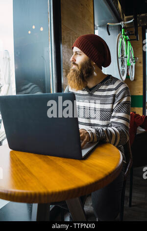 Hipster man looking through window while using laptop computer on table at cafe Stock Photo