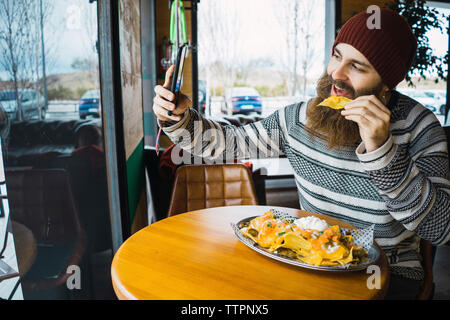 Hipster man taking selfie while eating tortilla chip with smart phone at cafe Stock Photo