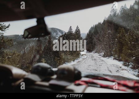 Snow covered road by trees in forest seen through rear windshield Stock Photo