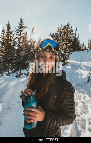Portrait of smiling young woman wearing ski goggles while holding water bottle on snowy field Stock Photo