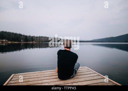 Rear view of man looking at calm lake while sitting on pier against cloudy sky in forest during sunset Stock Photo