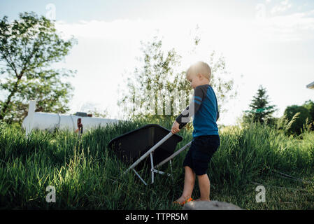 Side view of boy holding wheelbarrow while standing on grassy field against sky Stock Photo