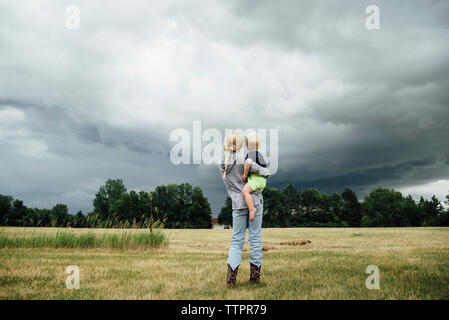 Rear view of sister carrying brother while standing on grassy field against cloudy sky Stock Photo