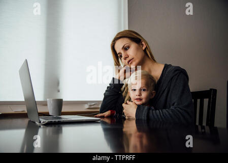 Portrait of baby boy sitting with mother using laptop on table at home Stock Photo