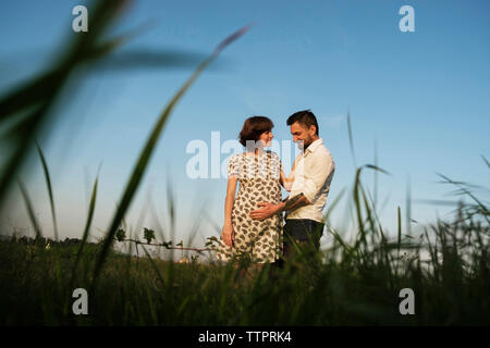 Low angle view of man touching PRegnant woman's stomach while standing on grassy field against clear sky Stock Photo