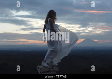 Full length of young woman wearing white dress while standing on pedestal against cloudy sky during sunset