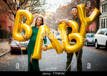 Portrait of happy expectant couple holding helium baby text balloons while standing on road in city Stock Photo